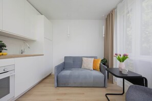 A modern, compact living space with a kitchenette, a grey sofa with cushions, a small table with a vase of flowers, and a window with curtains. - Functional Studio near Pole Mokotowskie Workspace Smart TV (Warszawa)