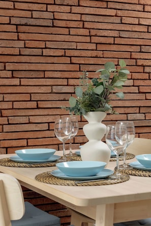 Ein Tisch ist gedeckt mit blauen Tellern, Weingläsern und einer weißen Vase mit Grünpflanzen vor einer Ziegelwand.
