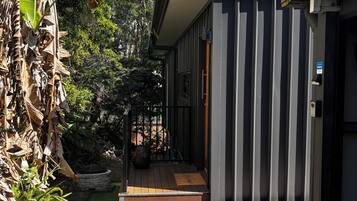 Un edificio moderno con terraza de madera, maceta de piedra y un jardín bien cuidado con césped artificial y palmeras.