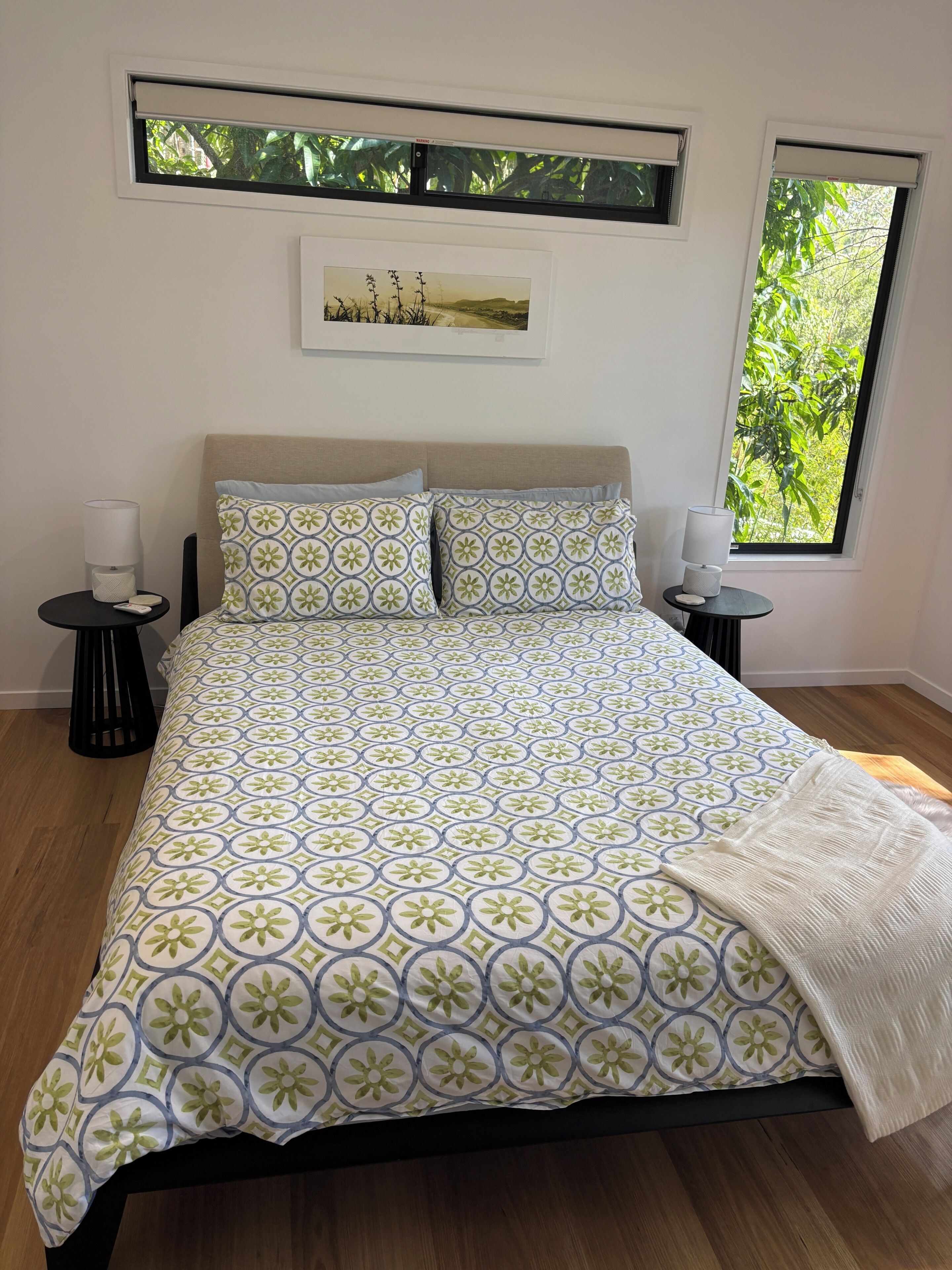 A bed with a patterned bedspread, two bedside tables with lamps, a window with a view of greenery, and a framed picture on the wall.