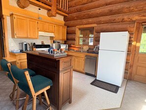 A wooden cabin interior with a kitchen area, wooden bar stools, and a refrigerator. - No-Ferry Camano Log Cabin w/ Puget Sound Views, Beach Access & EV Charger (Camano)