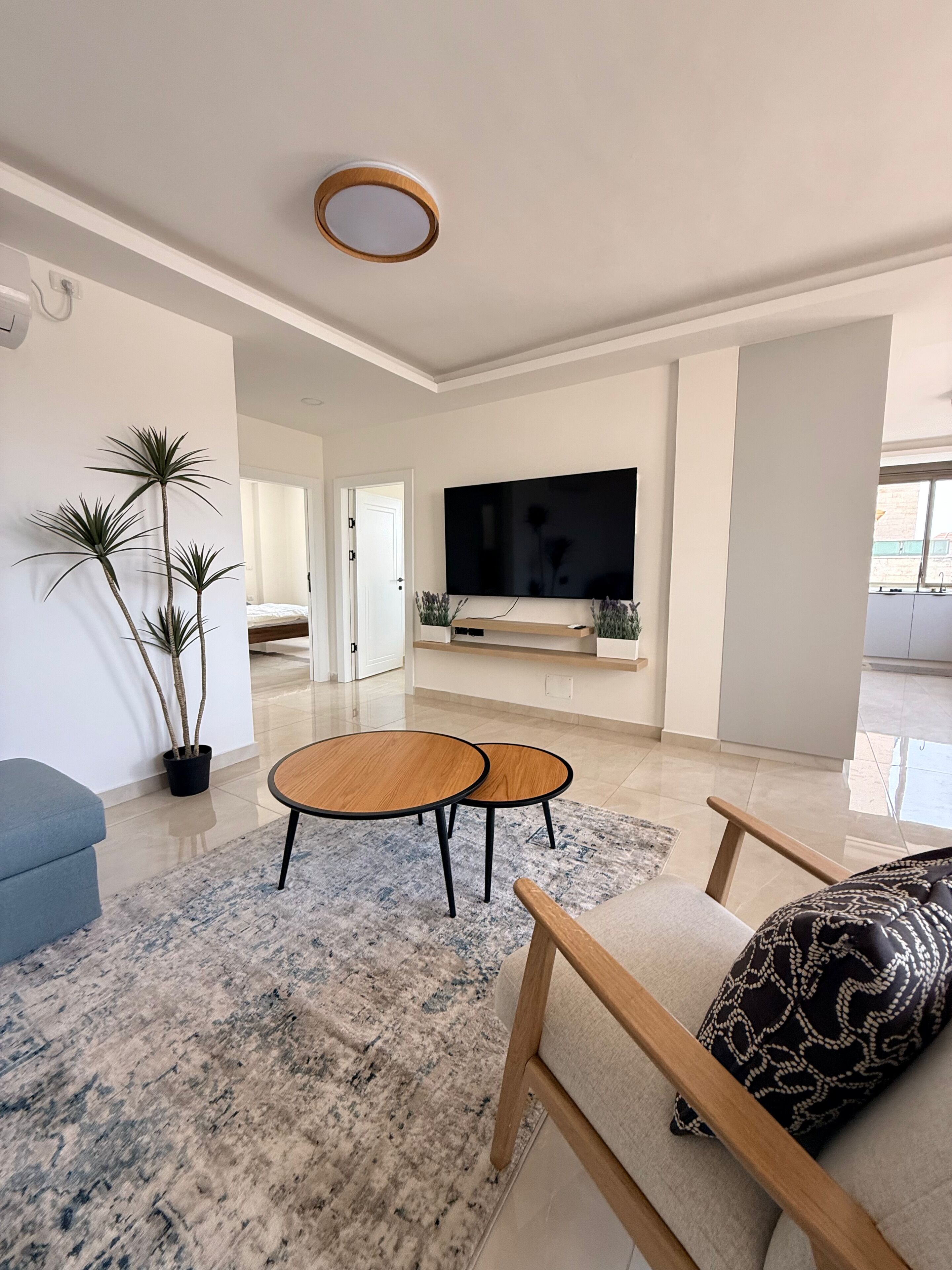 A modern living room with a flat-screen TV, two round wooden tables, a blue sofa, and a patterned throw pillow.