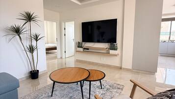 A modern living room with a flat-screen TV, two round wooden tables, a blue sofa, and a patterned throw pillow.