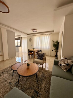 A modern living room with a sofa, two coffee tables, a dining table with chairs, and a large window with curtains.