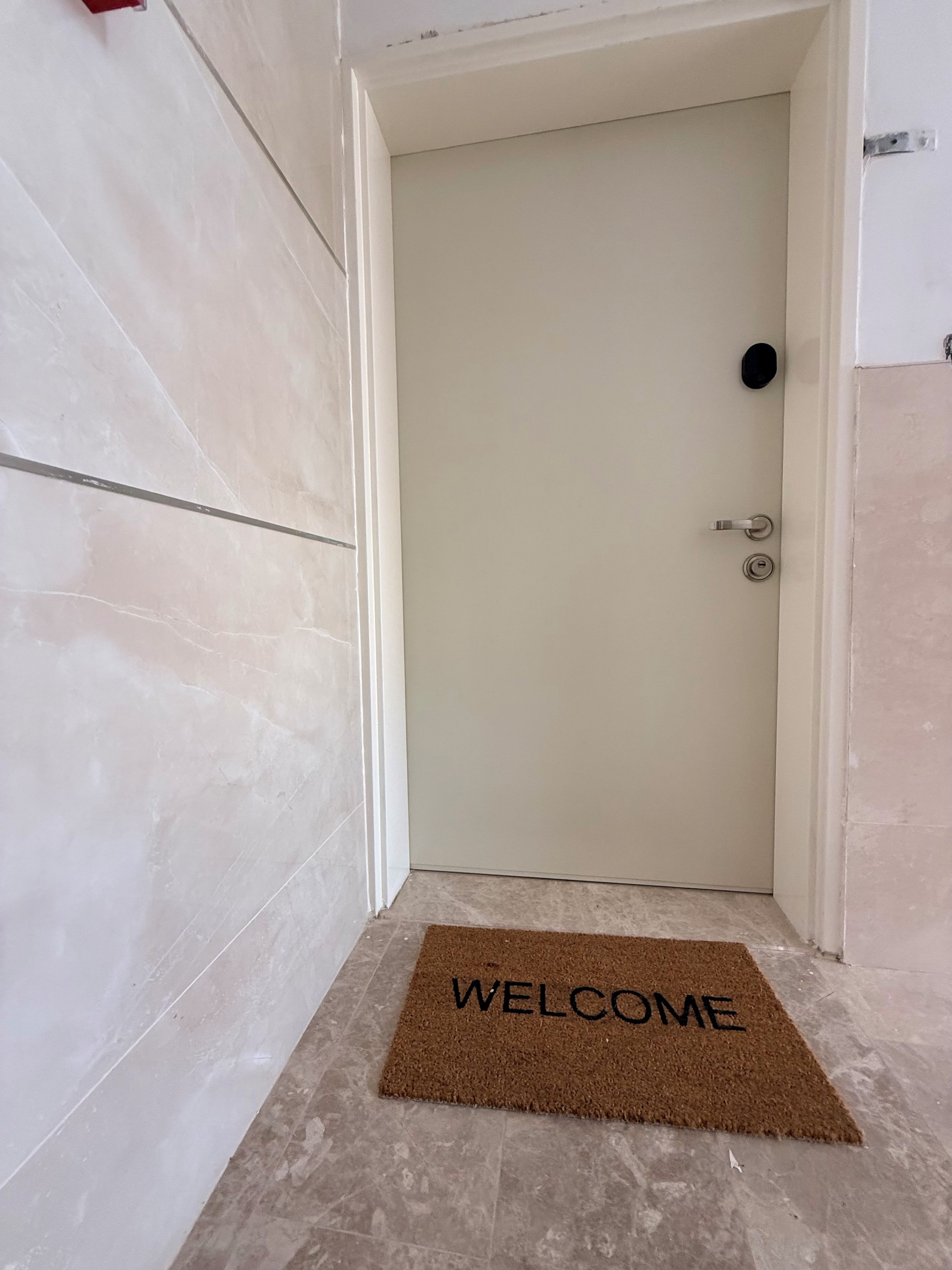 Une porte avec une poignée de porte et un paillasson sur lequel est écrit « BIENVENUE », le tout posé sur un sol carrelé.