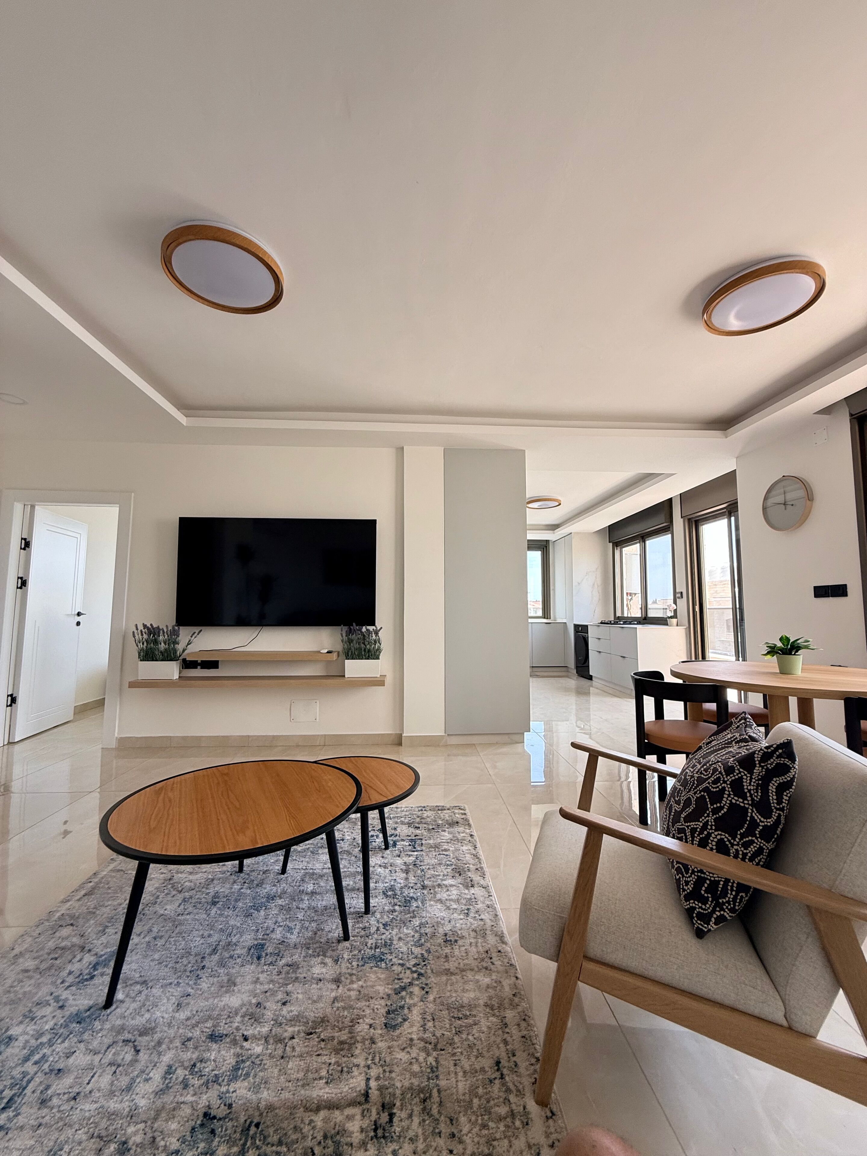 A modern living room with a flat-screen TV, two round wooden tables, a sofa with patterned cushions, and a large window providing natural light.
