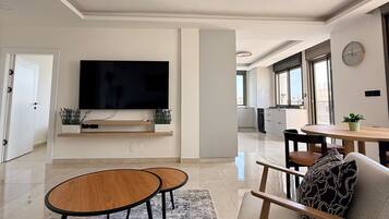 A modern living room with a flat-screen TV, two round wooden tables, a sofa with patterned cushions, and a large window providing natural light.