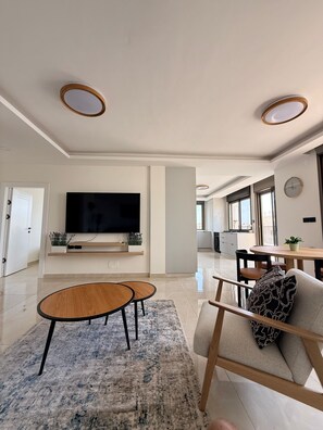 A modern living room with a flat-screen TV, two round wooden tables, a sofa with patterned cushions, and a large window providing natural light. - 3-bedroom apartment in beautiful Masyon in Ramallah (Ramallah)