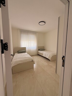 Ein Schlafzimmer mit zwei Betten, einem Fenster mit Jalousien, einer Tür und einer Leuchte.