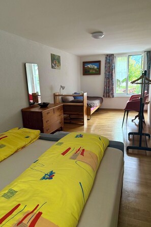 Quarto com duas camas, penteadeira, escrivaninha, cadeira, cabideiro e janela com vista para o verde.