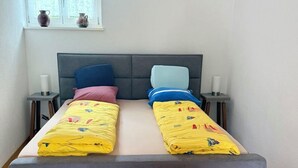 Een tweepersoonsbed met een grijs hoofdbord, twee nachtkastjes met lampen, een bankje aan het voeteneind van het bed en een raam met gordijnen.