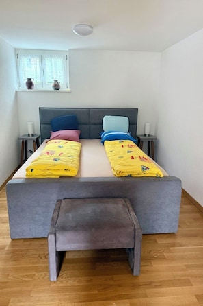 Ein Doppelbett mit grauem Kopfteil, zwei Nachttische mit Lampen, eine Sitzbank am Fußende des Bettes und ein Fenster mit Vorhängen.