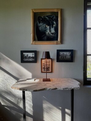 A room with a framed artwork of a cat's face, two smaller framed pictures, a table with a lamp, and a window with natural light.