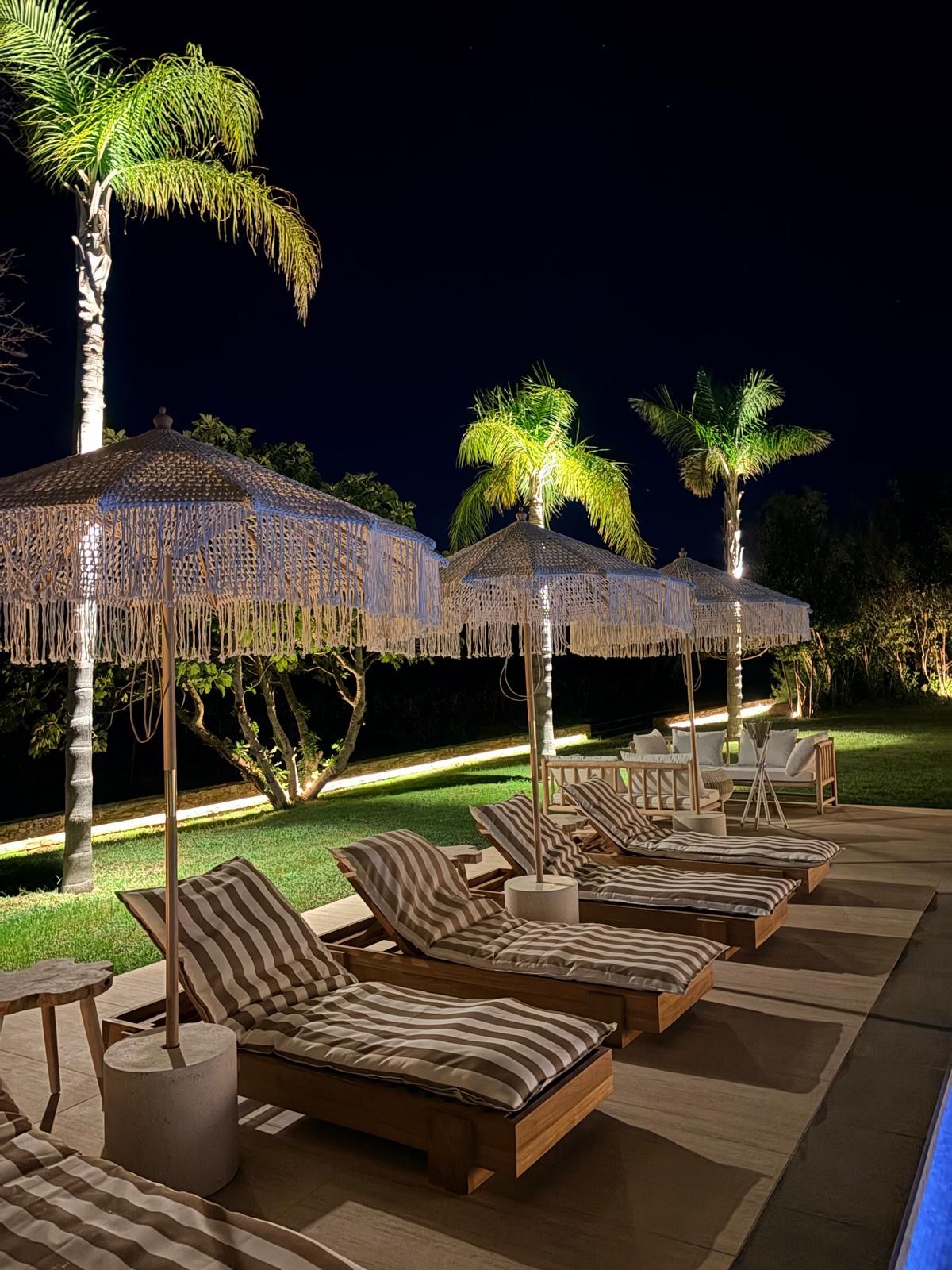 Un espace de détente extérieur aménagé pour la nuit, avec des chaises longues à rayures et des parasols en chaume, le tout entouré de palmiers.