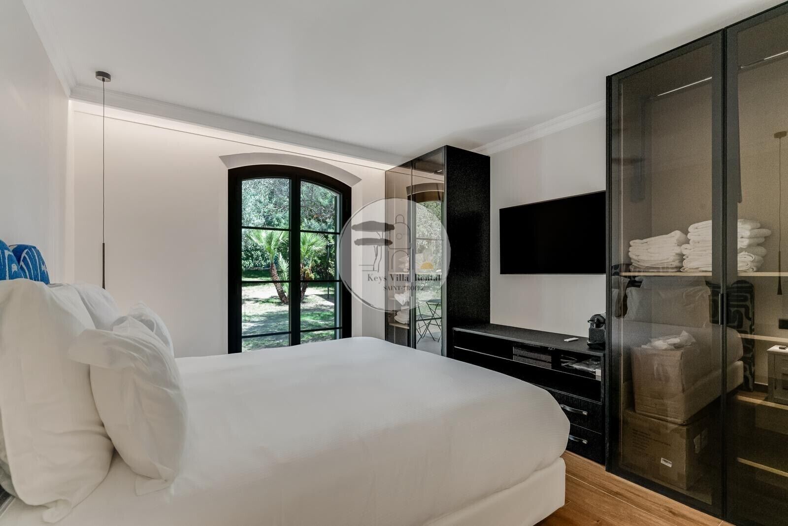 A modern bedroom with a large bed, a TV mounted on the wall, and a glass cabinet with towels.