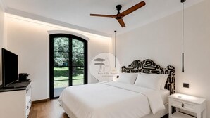 Ein modernes Schlafzimmer mit einem großen Bett, einem Deckenventilator, einem Fernseher und Blick auf einen Garten durch eine Glastür.