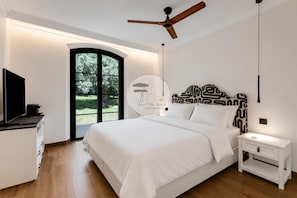 Ein modernes Schlafzimmer mit einem großen Bett, einem Deckenventilator, einem Fernseher und Blick auf einen Garten durch eine Glastür.