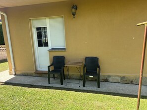 Un patio con dos sillas y una mesita frente a una pared amarilla, una puerta con persianas y una lámpara fijada a la pared.