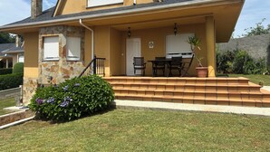 A two-story house with a stone facade, a front porch with a table and chairs, and a small garden with purple flowers. - Family home with a garden: Beach, mountains, and pets welcome (Garage included) (La Caridad)