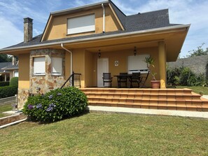 Una casa de dos pisos con fachada de piedra, un porche con mesa y sillas, y un pequeño jardín con flores moradas.
