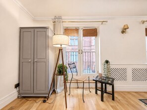 A room with a grey cabinet, a floor lamp, a small table with a plant, a chair, and a window with blinds. - Central Garden Townhouse near Oxford St and SoHo (London)