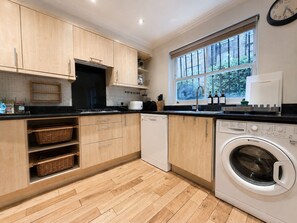 A modern kitchen with wooden cabinets, a built-in washing machine, and a window with a view of greenery. - Central Garden Townhouse near Oxford St and SoHo (London)