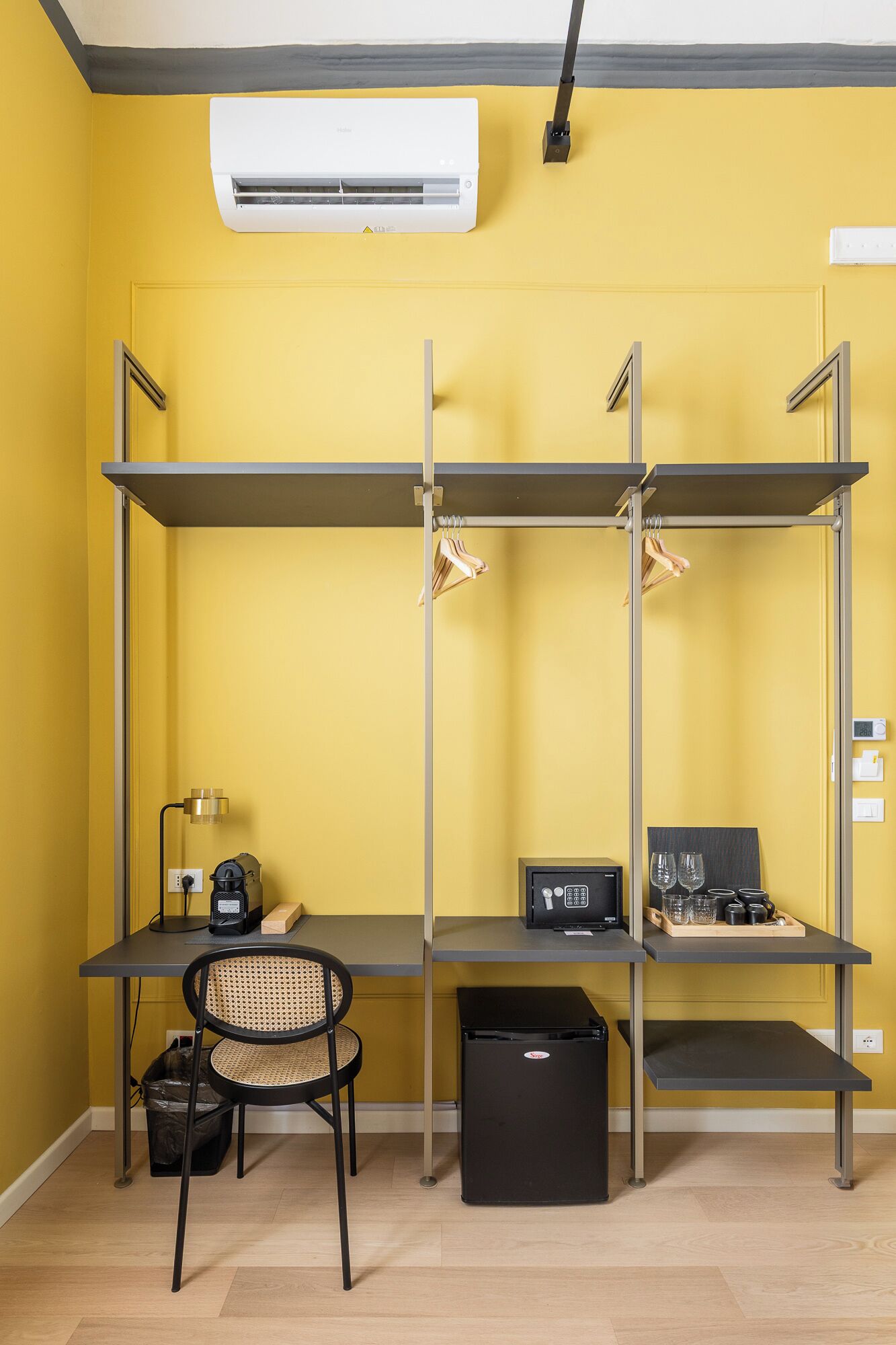 Een kamer met een gele muur, een bureau met een computer, een stoel, een prullenbak en een plank met kleding.