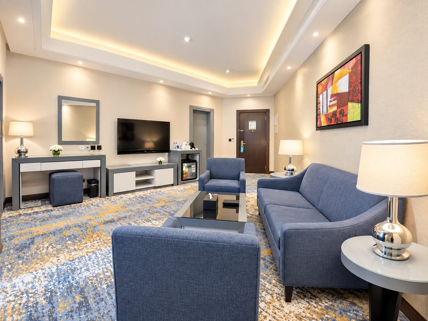 A hotel room with a flat-screen TV, a sofa, a chair, a desk with a lamp, and a coffee machine.