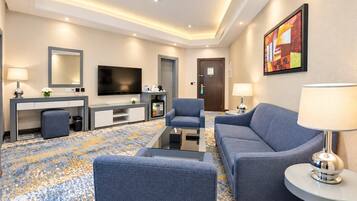 Habitación de hotel con televisión de pantalla plana, un sofá, una silla, un escritorio con lámpara y una cafetera.