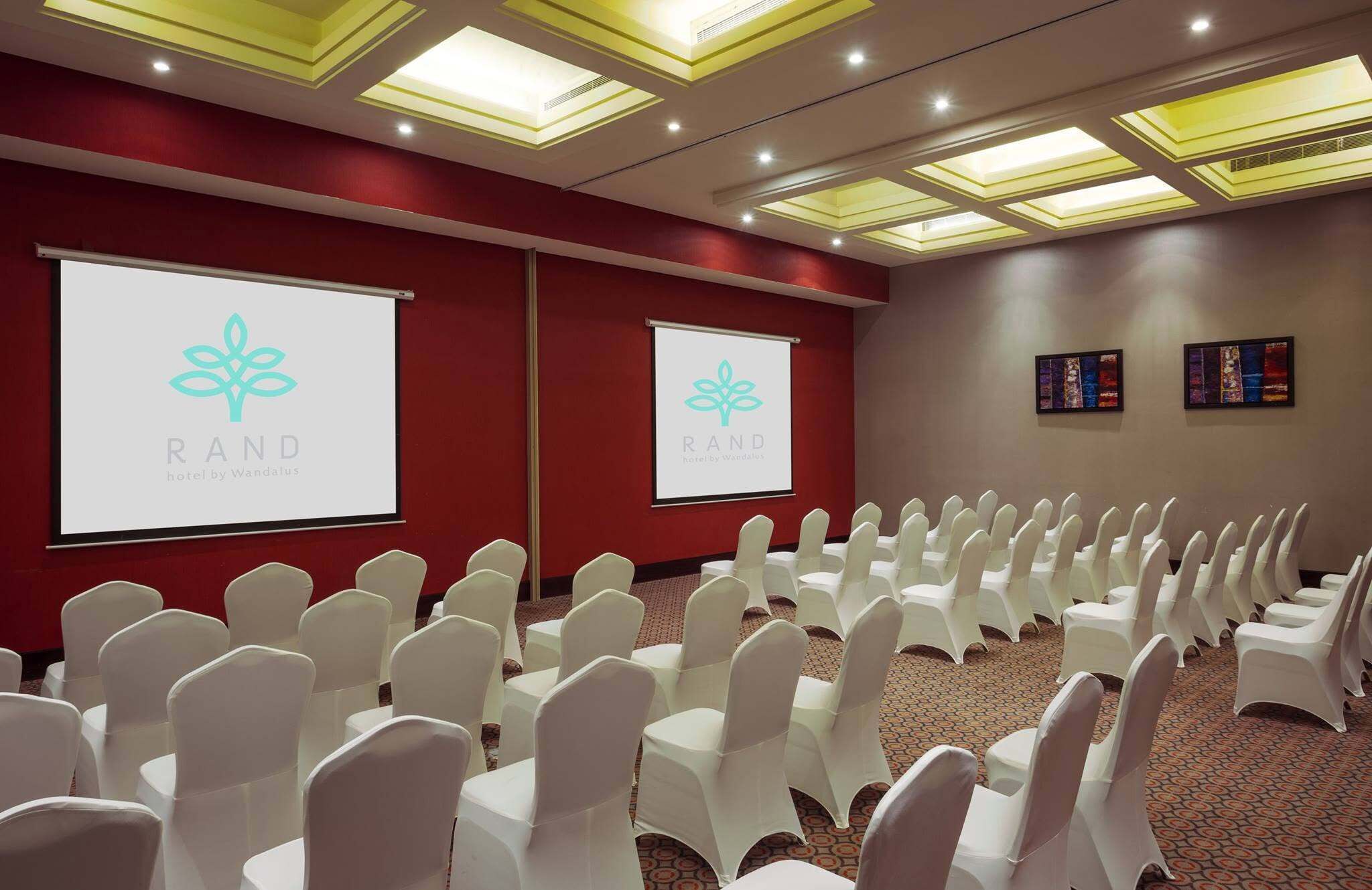 A conference room with rows of white chairs, a projector screen with the 'RAND' logo, and a carpeted floor.