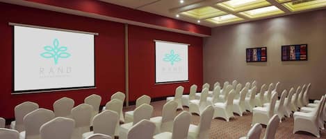 Un salón de conferencias con filas de sillas blancas, una pantalla de proyector con el logotipo de 'RAND' y suelo alfombrado.