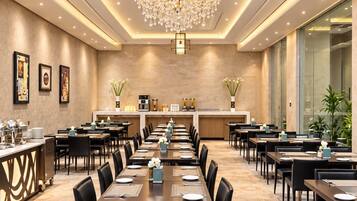A spacious hotel dining area with long tables, chairs, a central chandelier, and a buffet counter.