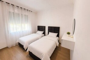 Ein Zimmer mit zwei Betten, einem Nachttisch, einem Spiegel und einem Fenster mit leichtem Vorhang.
