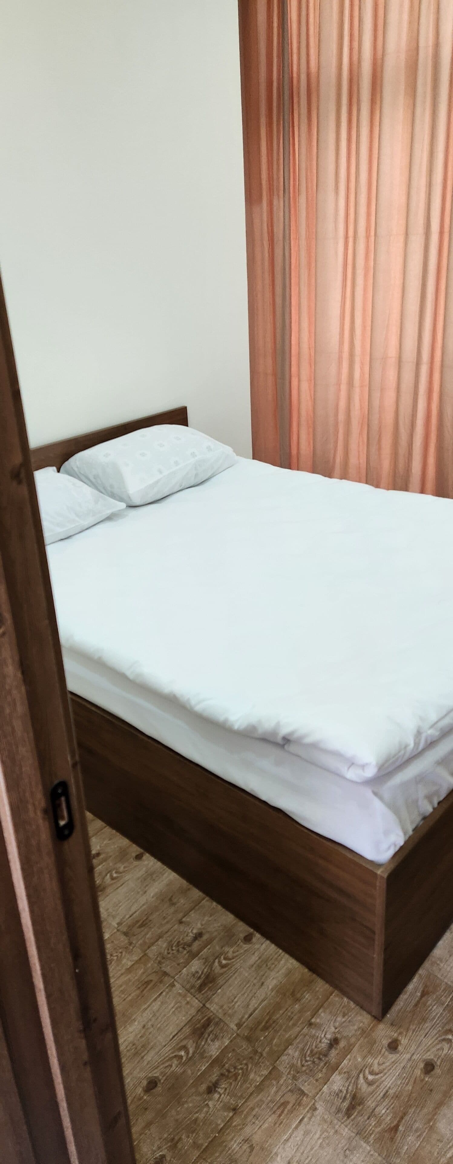 A single bed with a wooden frame and white bedding in a room with wooden flooring and a curtain.