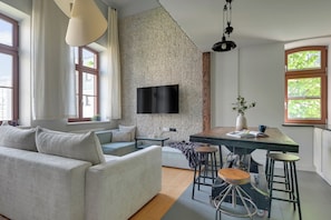 A modern living room with a sofa, a dining table with stools, a flat-screen TV mounted on a textured wall, and large windows.