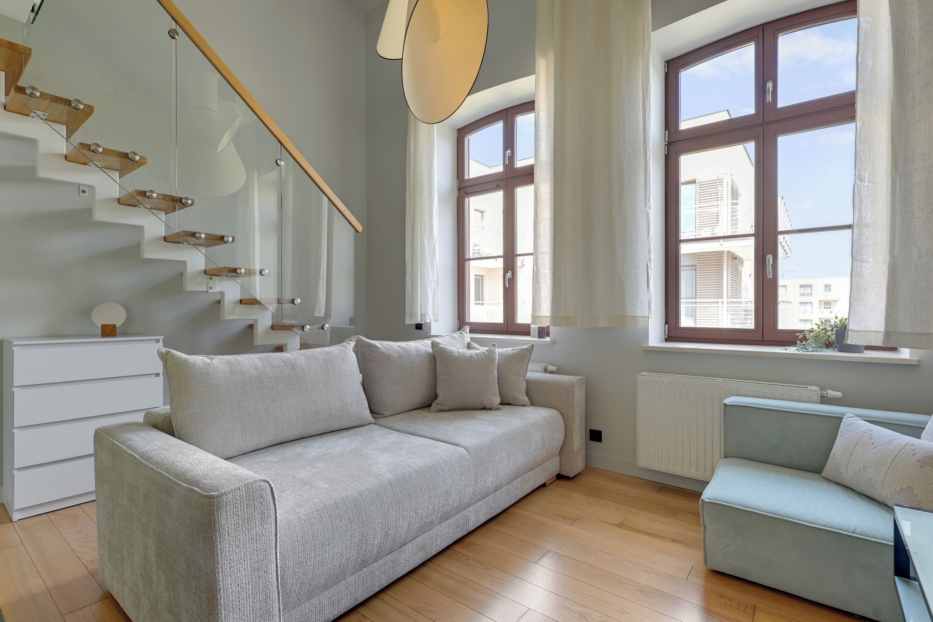 Una sala de estar moderna con un sofá, un sillón más pequeño, una escalera y ventanas amplias.