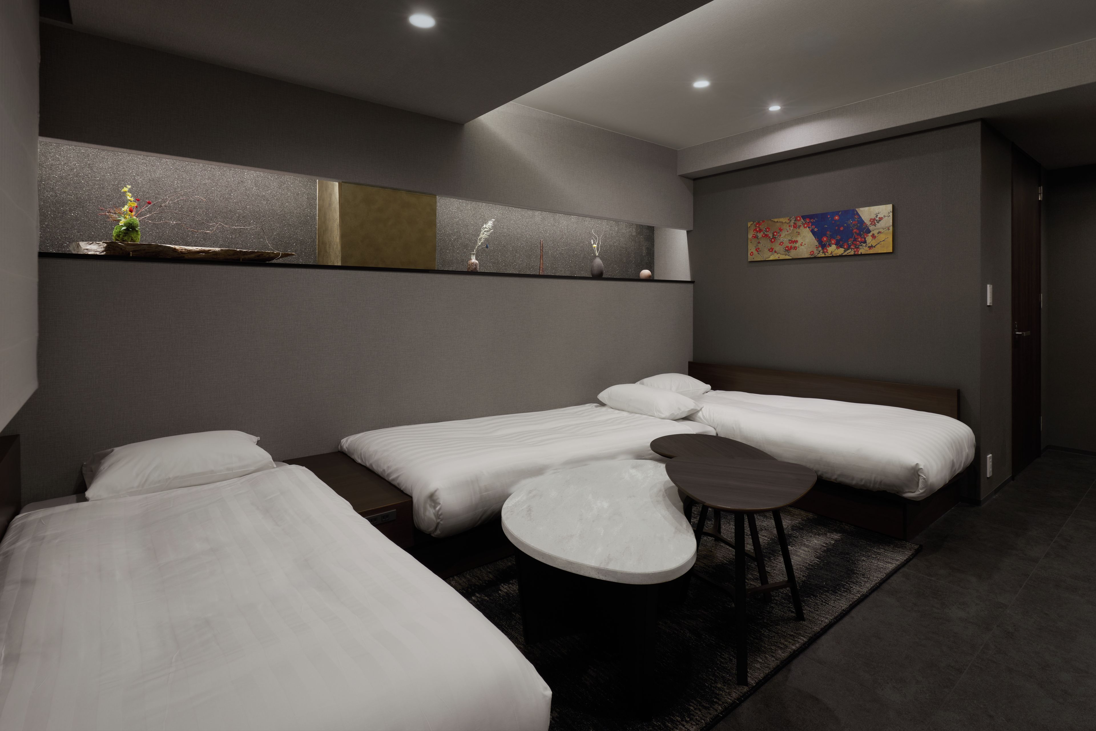 A modern hotel room with three beds, a small round table, and a painting on the wall.
