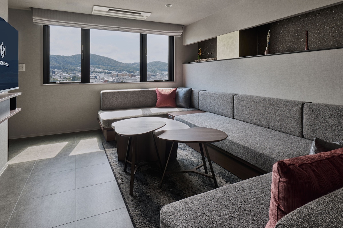 A modern living room with a grey sectional sofa, two round tables, and a large window offering a city view.