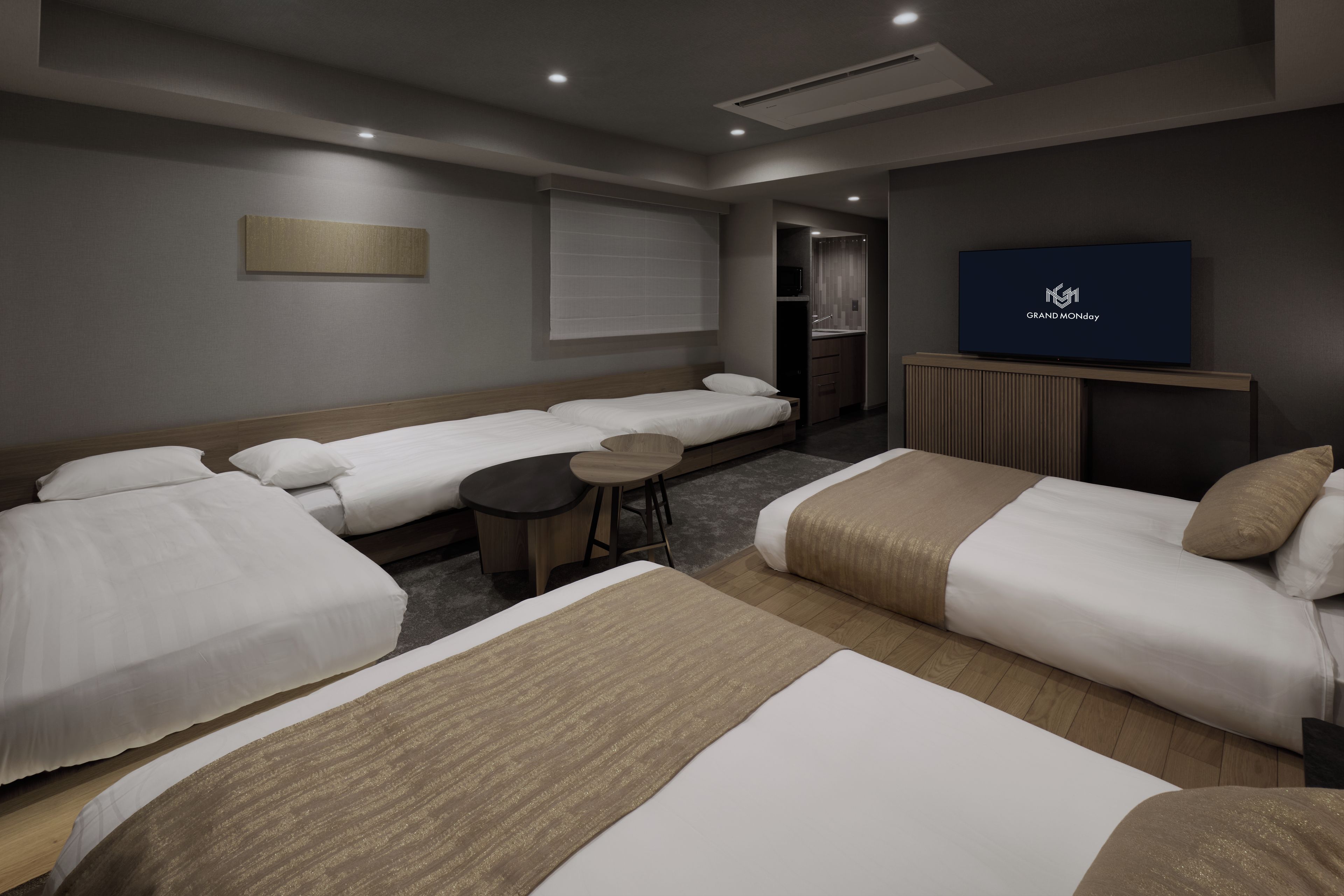 A hotel room with two single beds, a sofa bed, a small round table, a television, and a bathroom with a mirror and sink.