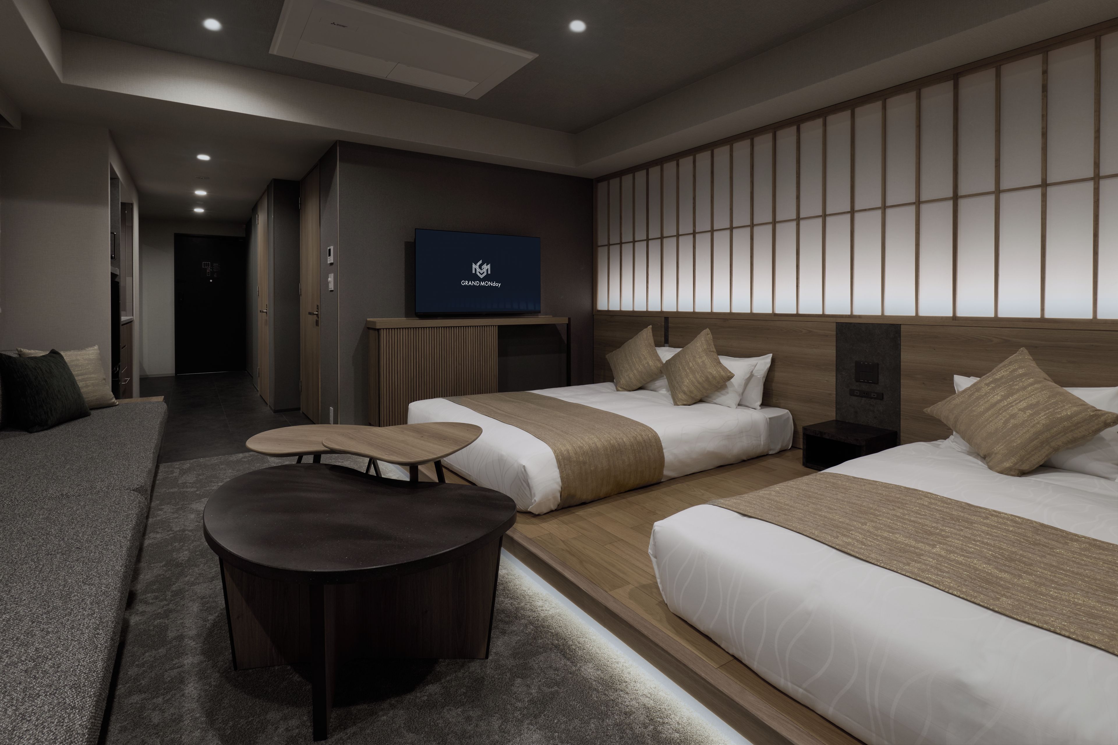 Una habitación de hotel moderna con un sofá, dos camas, una mesita y un televisor mostrando 'GRAND MONDAY'.