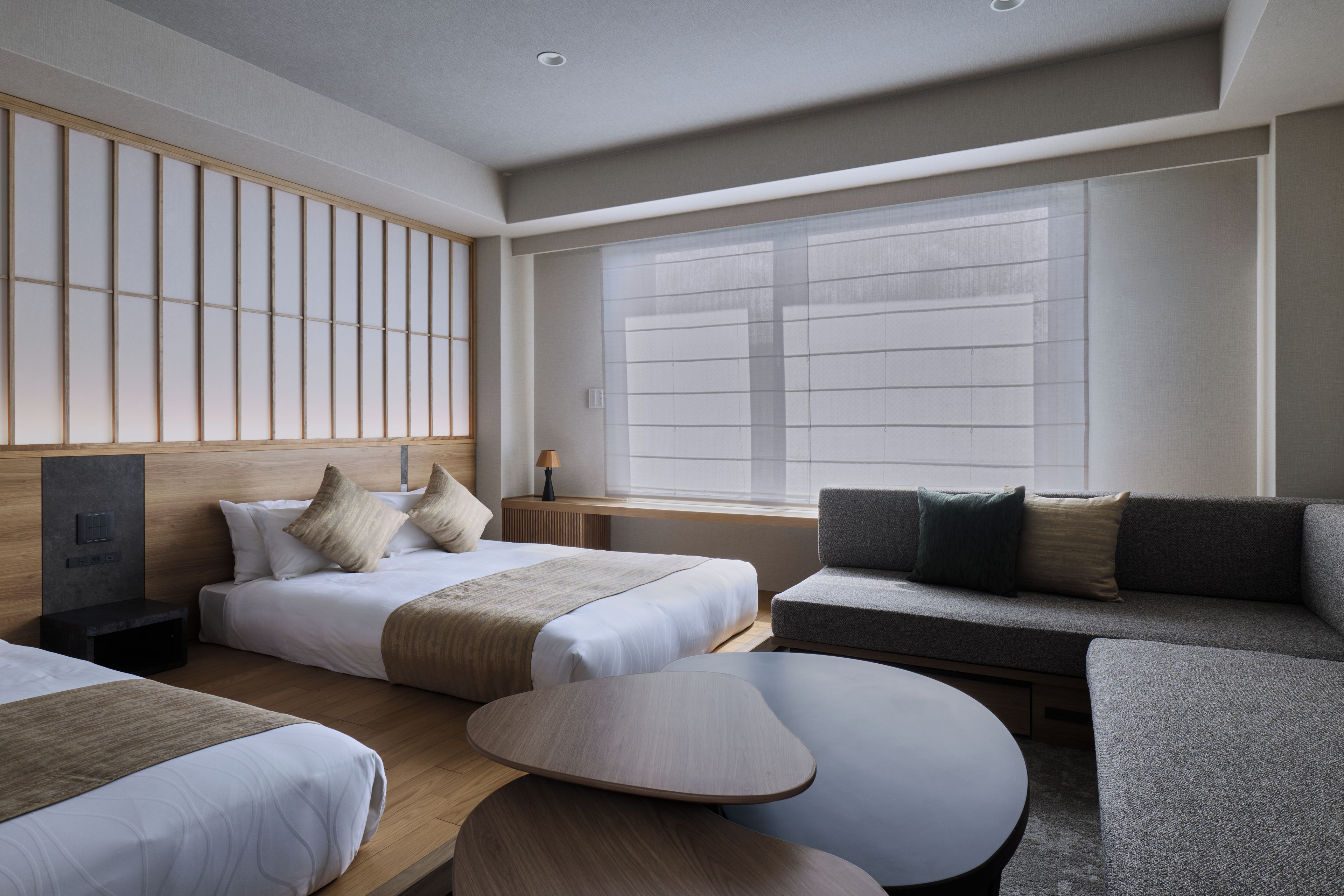 Quarto de hotel moderno com duas camas, um sofá, uma mesinha e uma janela grande.