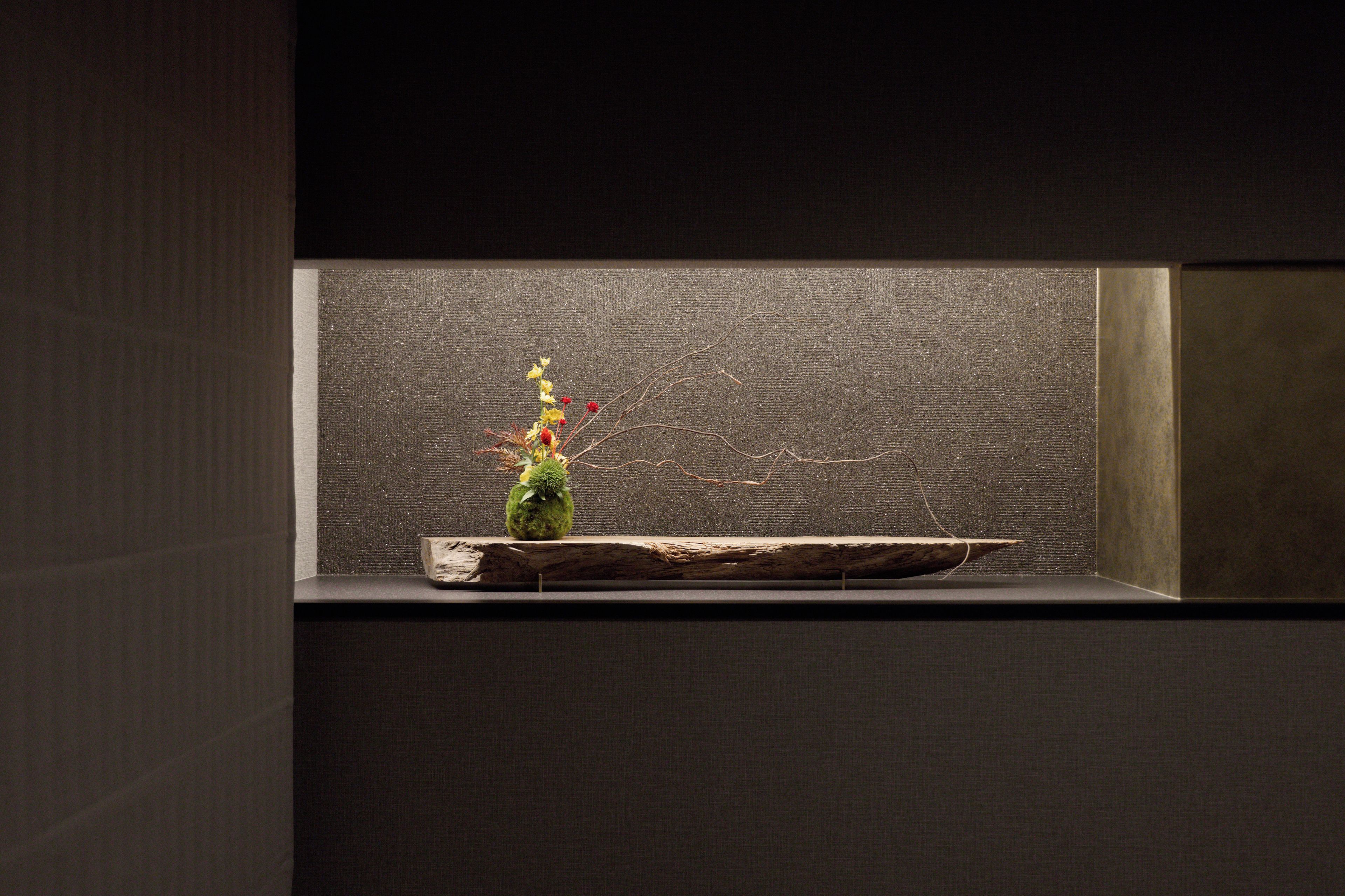 Una habitación oscura con pared texturizada y un estante de madera que sostiene un jarrón pequeño con flores secas.