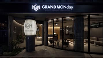 The exterior of a hotel with a modern facade, a prominent illuminated logo, and a reception area visible through the glass doors.