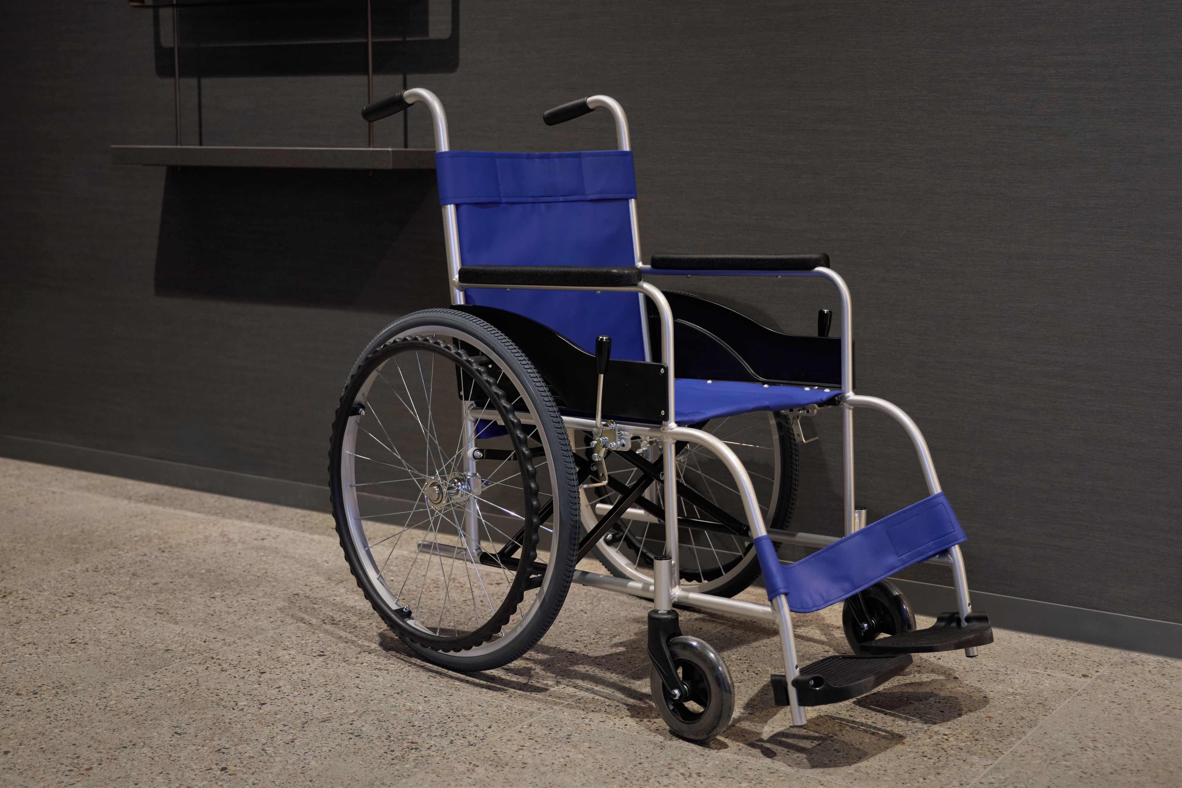 Una silla de ruedas con asiento y respaldo azules, sobre un piso de concreto frente a una pared oscura.