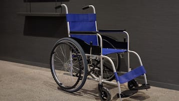 En rullstol med ett blått säte och ryggstöd, placerad på ett betonggolv framför en mörk vägg.