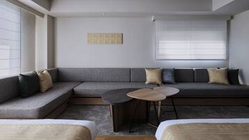 Une pièce moderne et minimaliste, avec un canapé d’angle, une petite table ronde et une fenêtre équipée de stores.