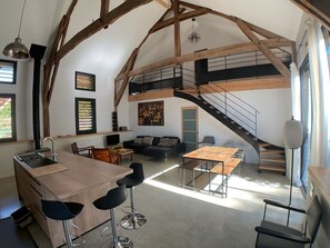 A spacious loft with a high ceiling, exposed wooden beams, a central staircase, a kitchen area with a bar, and a seating area with a sofa and chairs. - Vacation Rental with Pool for 6 People in the Heart of the Périgord Noir (Veyrines-de-Domme)