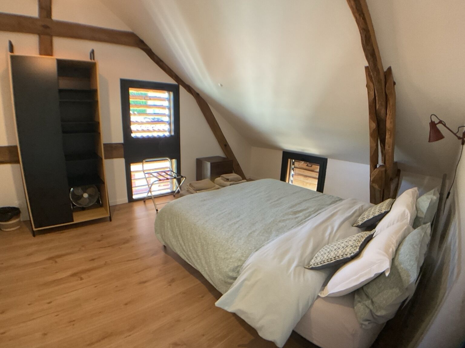 A bedroom with a sloped ceiling, a bed with pillows, a wooden floor, a built-in wardrobe, and a window with blinds.