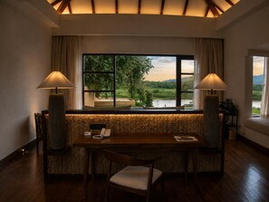 A dining area with a view of a river and hills, featuring a long table, chairs, and two lamps. - The Postcard on the Rapti River (Meghauli)