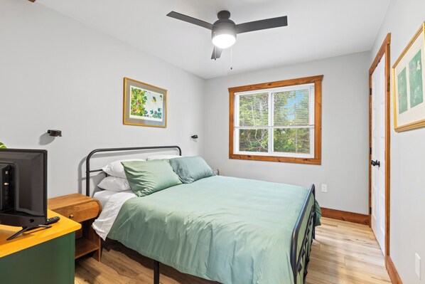 Ein Schlafzimmer mit einem Bett, einem Schreibtisch mit Computer, einem Deckenventilator, einem Fenster mit Jalousien und gerahmten Kunstwerken an den Wänden.
